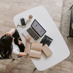 From above of young woman with long dark hair in casual clothes working at table and browsing netbook while sitting in modern workplace and touching hair