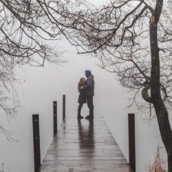 man and woman facing each other and standing on beach dock surrounded with bare trees under foggy season