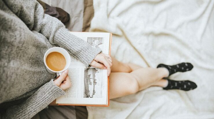 woman holding a cup of coffee at right hand and reading book on her lap while holding it open with her left hand in a well-lit room